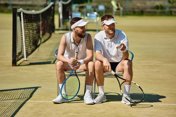 Two men share laughter and drinks while resting on a tennis court during summer play. — Stock Photo