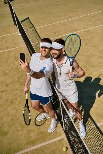 Dos hombres felizmente se toman una selfie juntos después de un divertido juego de tenis a la luz del sol de verano. - foto de stock