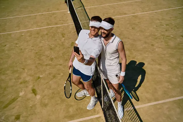 Dos jóvenes sonríen y se toman una selfie juntos en una soleada cancha de tenis durante el verano. - foto de stock
