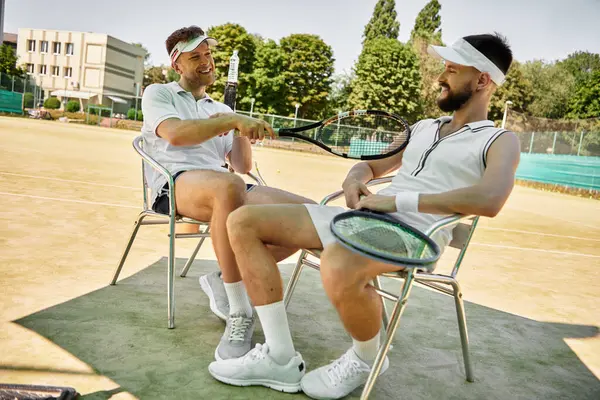Two men relax in chairs on the tennis court, sharing laughter after a fun game. — Stock Photo