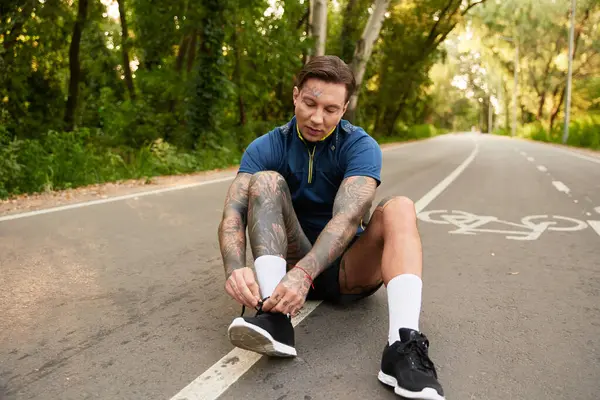 Handsome young man pauses during a walk to tie his shoes on a quiet street surrounded by trees. — Stock Photo