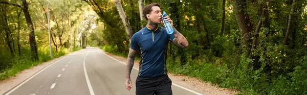 Handsome young man strolls along a serene path, sipping water amidst lush green surroundings. — Stock Photo