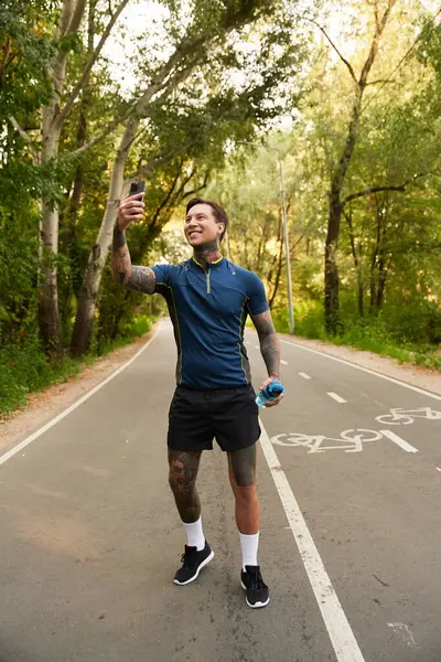 A handsome young man with tattoos is capturing a selfie while walking on a tree lined path. — Stock Photo