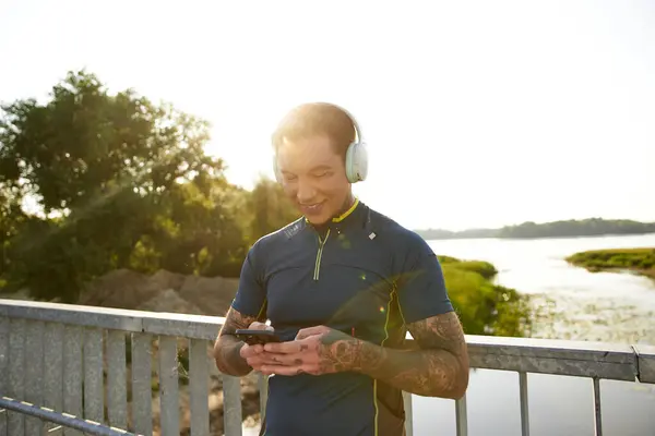 Um homem jovem e atraente caminha ao longo de uma margem do rio, imerso em música, sorrindo para o telefone. — Fotografia de Stock