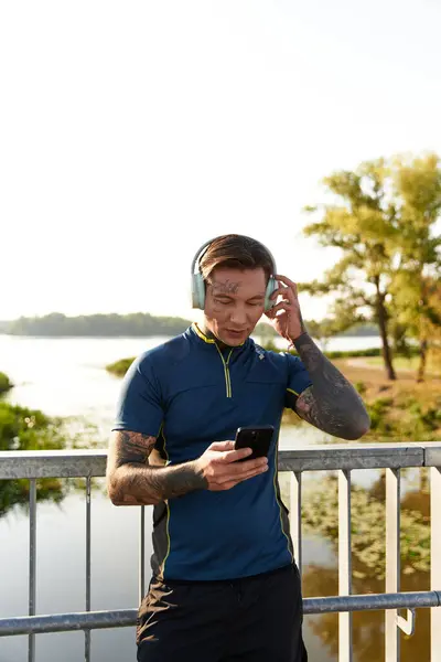A young man strolls along a scenic riverside, engaged with his smartphone and headphones. — Stock Photo