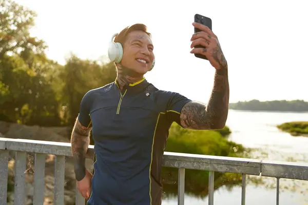Ein tätowierter junger Mann hält auf einem Spaziergang inne und lächelt für ein Natur-Hintergrund-Selfie. — Stockfoto
