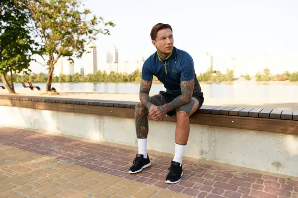 A young man with tattoos relaxes on a bench while taking a break from his walk along the river. — Stock Photo