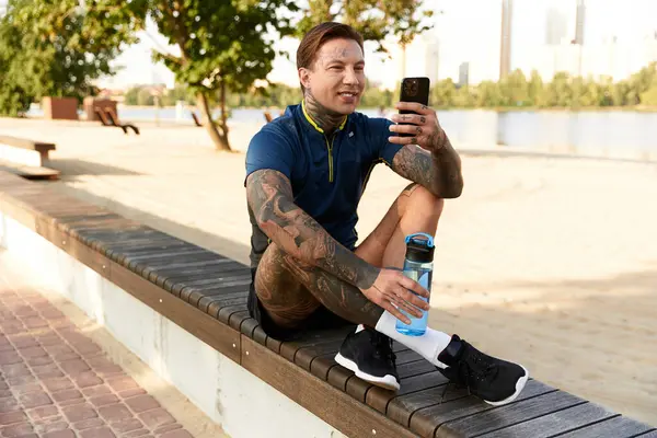 Handsome young man sits on a bench by the lake, smiling while using his smartphone. — Stock Photo