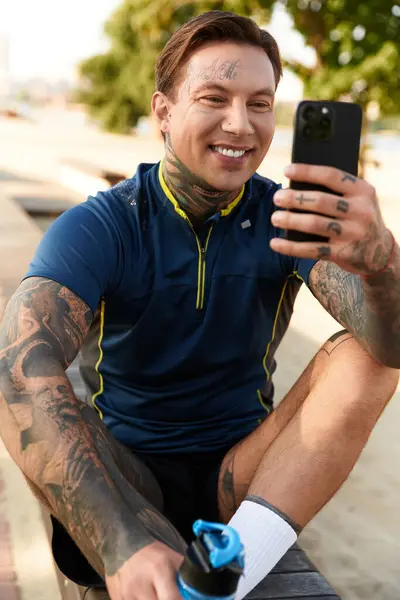 A young man with tattoos relaxes on a bench, smiling as he checks his phone during a walk. — Stock Photo