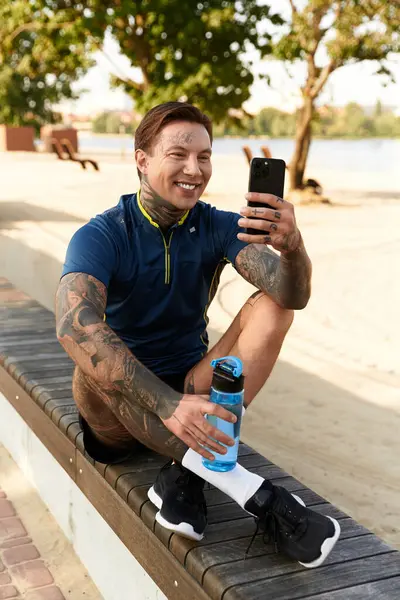 Niño con tatuajes sonríe en su teléfono, tomando un descanso en un sendero de jogging junto al lago soleado. - foto de stock