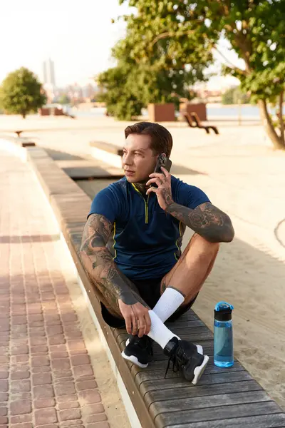 A young man with tattoos takes a break on a bench, making a phone call and enjoying the sun. — Stock Photo