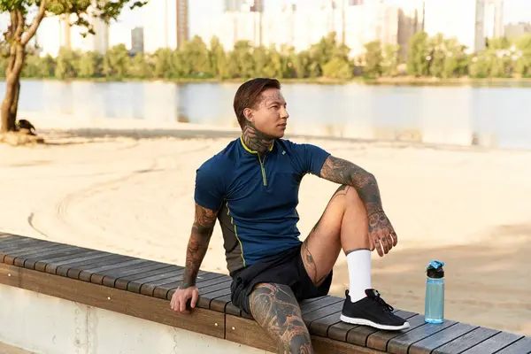 A handsome young man with tattoos pauses on a bench, taking in the serene riverside atmosphere. — Stock Photo