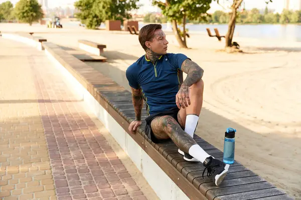 A young man with tattoos takes a break on a bench during his scenic walk by the river. — Stock Photo