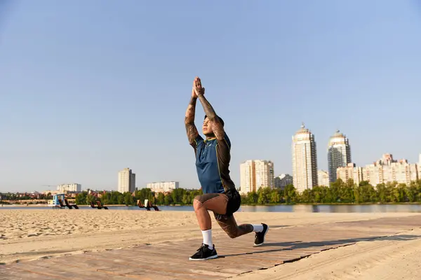 A handsome young man does yoga on the sunny beach with city buildings behind him. — Stock Photo