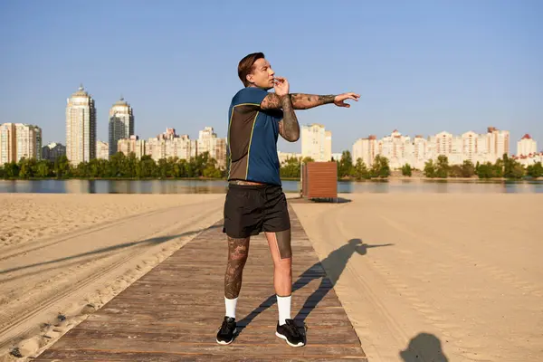 A young man stretches his arms while walking peacefully by the river, amid tall buildings and trees. — Stock Photo