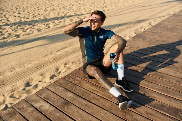 Un joven guapo está tomando un descanso de su paseo, sentado en un paseo marítimo de madera junto a la playa. - foto de stock