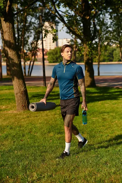 A young man confidently walks through a sunny park with a yoga mat and water bottle. — Stock Photo