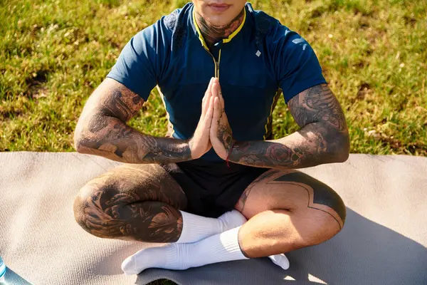 Joven guapo se sienta con las piernas cruzadas en una alfombra, meditando en la naturaleza rodeada de vegetación. - foto de stock