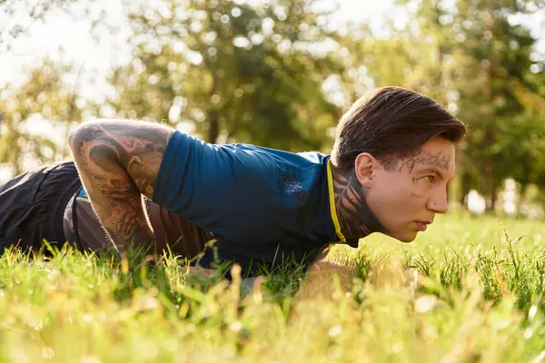 Um jovem com tatuagens realiza flexões na grama em um parque iluminado pelo sol. — Fotografia de Stock