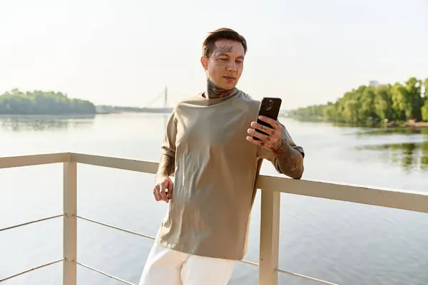 A young man stands on a riverside balcony, admiring the view and using his phone under sunlight. — Stock Photo