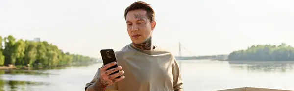 Young man strolls along a picturesque riverside, absorbed in his phone as the sun shines bright. — Stock Photo