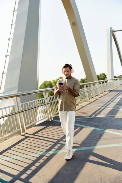 A handsome young man strolls along a bridge, absorbed in his phone during a sunny afternoon. — Stock Photo