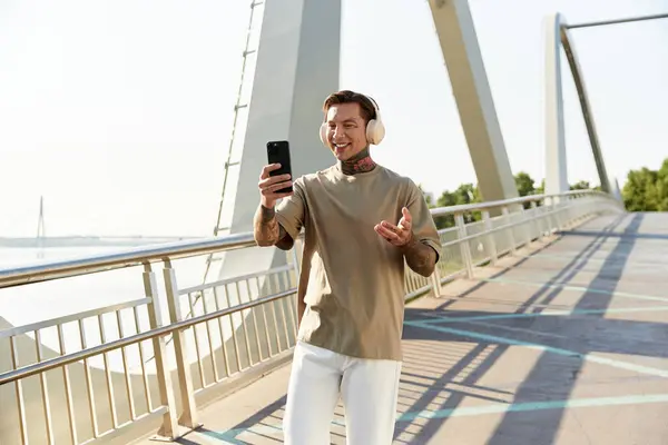 A stylish young man smiles while walking along a scenic river walkway, wearing headphones. — Stock Photo