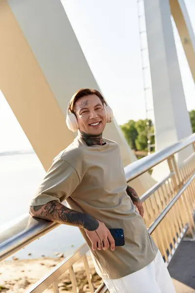 Jovem com fones de ouvido elegantes sorri enquanto relaxa em uma ponte com vista para a água. — Fotografia de Stock
