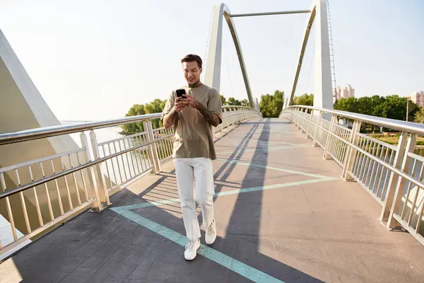 Young man strolls confidently on a bridge, smiling and scrolling through his smartphone. — Stock Photo