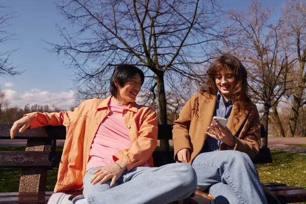 A young asian man and woman laugh together while sharing a phone in a sunny park. — Stock Photo