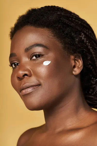 A young Black woman highlights her radiant skin with beauty products against a warm backdrop. — Stock Photo