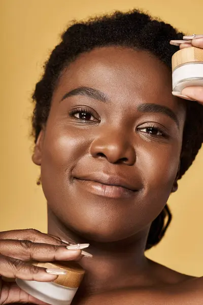 A young African American woman showcases her beautiful skin while holding skincare products. — Stock Photo