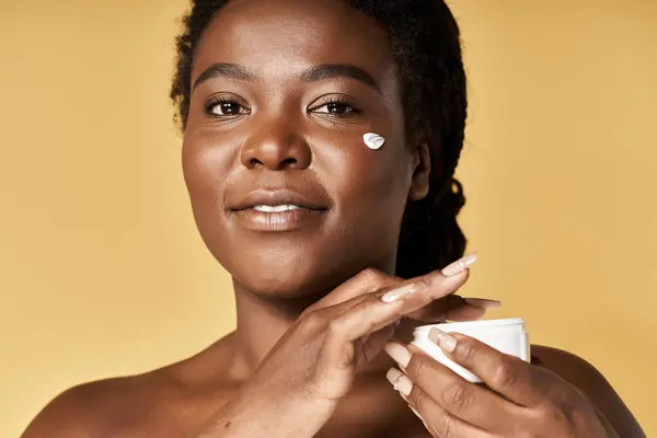 Jeune femme afro-américaine applique de la crème sur sa peau éclatante, rayonnant de confiance et de beauté. — Photo de stock