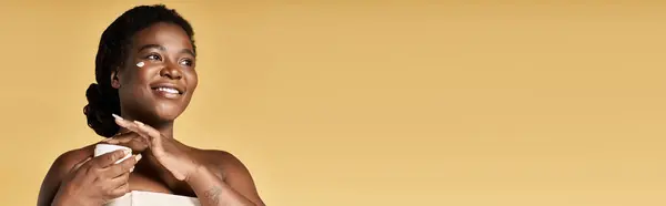 Uma jovem afro-americana aplicando produtos de cuidados com a pele em um espaço de estúdio, irradiando beleza, bandeira — Fotografia de Stock