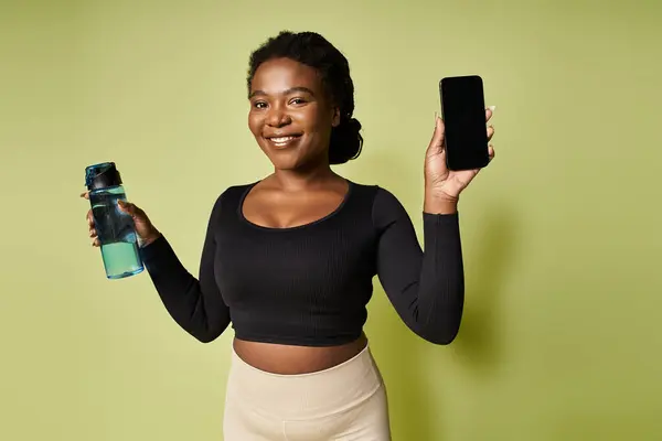 A fit young african american woman holds a water bottle and phone, poised for her exercise session. — Stock Photo