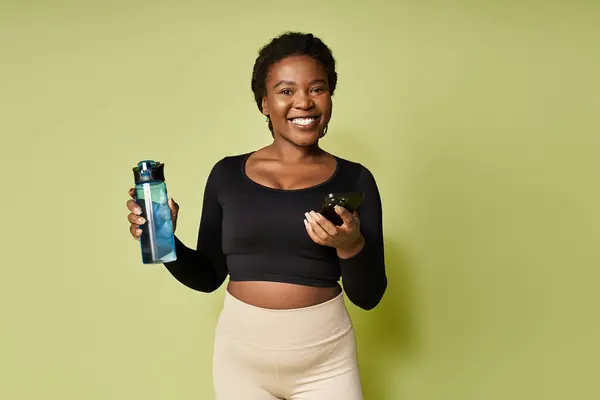 Sporty young woman holds a water bottle and phone, smiling confidently in a vibrant studio. — Stock Photo