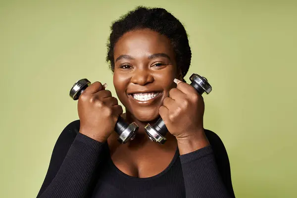 Desportista afro-americana energética levanta halteres com um sorriso radiante em um estúdio vibrante. — Fotografia de Stock