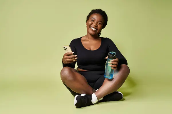 Young African American woman smiles while sitting on the floor, holding a water bottle and phone. — Stock Photo