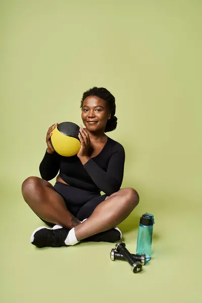 Fit young African American woman sits confidently, holding a medicine ball in a bright setting. — Stock Photo