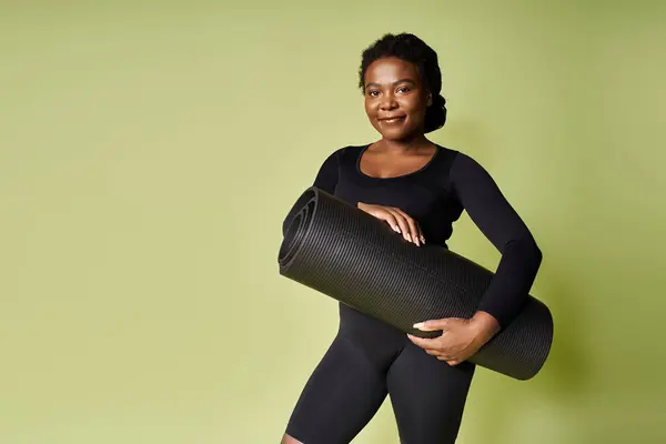 Una joven negra en forma se para con una esterilla de entrenamiento, lista para hacer ejercicio en un estudio moderno. - foto de stock