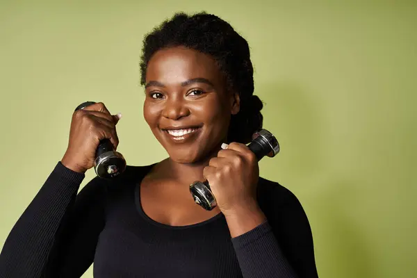 A young african american sportswoman smiles confidently while lifting dumbbells in a vibrant studio. — Stock Photo
