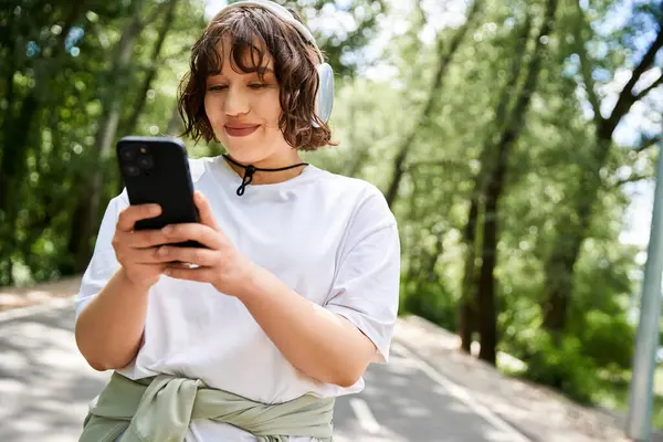 Una giovane donna sorride mentre usa il suo telefono in un ambiente verde e lussureggiante. — Foto stock