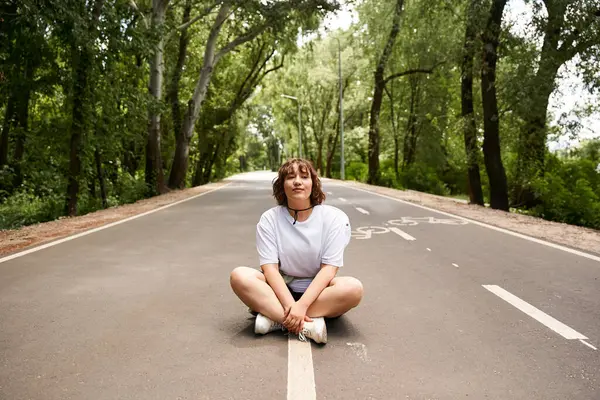 A young woman sits cross-legged on the road, soaking up the warm sun in a lush green setting. — Stock Photo