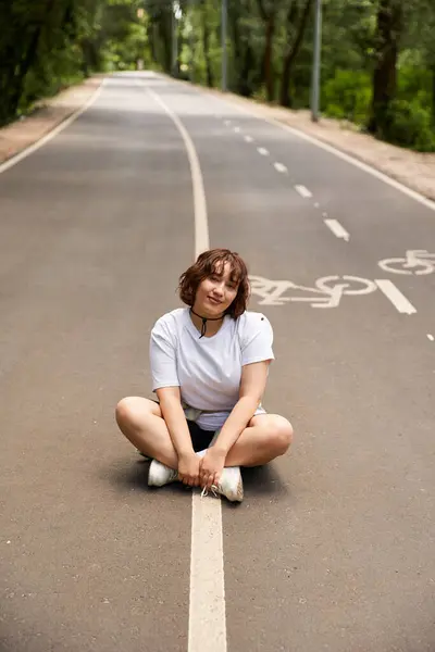 A young woman sits on a quiet road surrounded by trees, embracing the warmth of summer. — Stock Photo