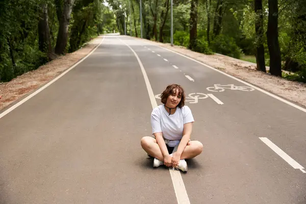 A cheerful young woman sits cross-legged on a serene pathway, soaking up the summer sun and nature. — Stock Photo