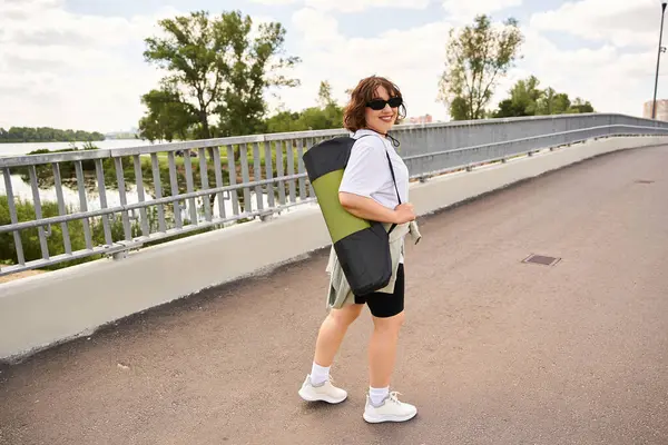 Fit young woman smiles as she carries her yoga mat along a scenic riverside pathway. — Stock Photo