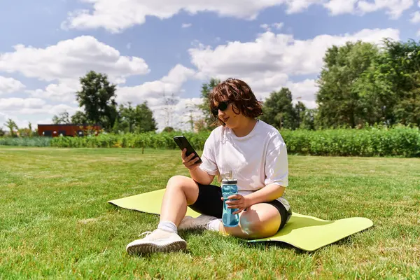 Fit woman relaxes on a mat outdoors, checking her phone and sipping water on a sunny day. — Stock Photo