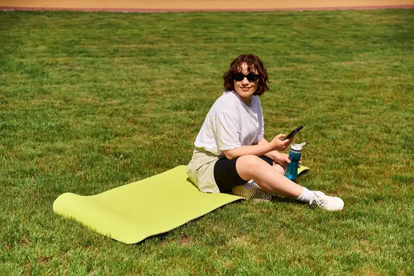 A fit woman takes a break on her mat while exercising in a vibrant green park under the sun. — Stock Photo