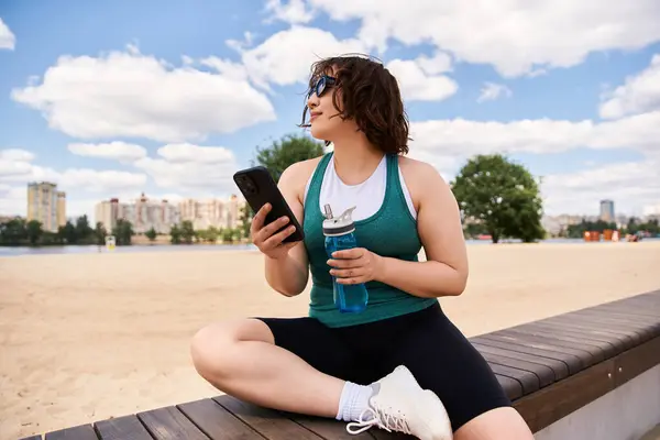 Una hermosa joven disfruta de un soleado día de verano sentada en un banco, mirando su teléfono. - foto de stock