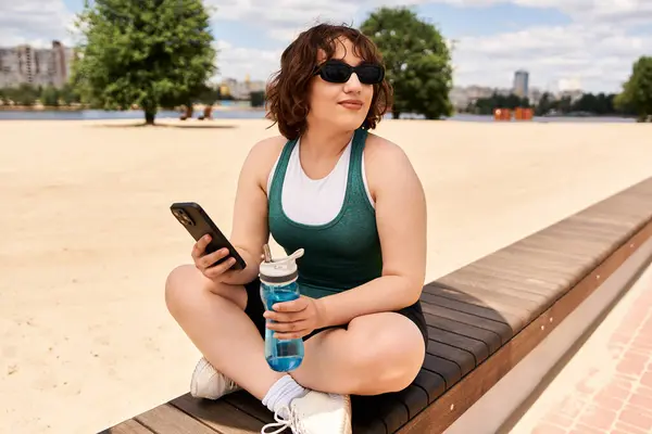 Bright summer day finds a young woman sitting outdoors, content with her phone and water bottle. — Stock Photo